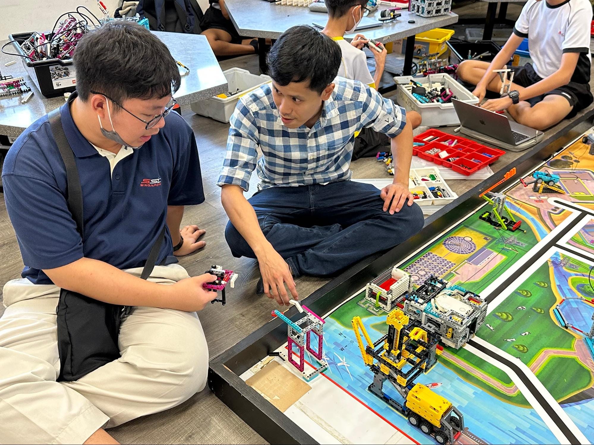 Two students sit on the floor working on a LEGO robotics competition mat with colorful brick constructions nearby.